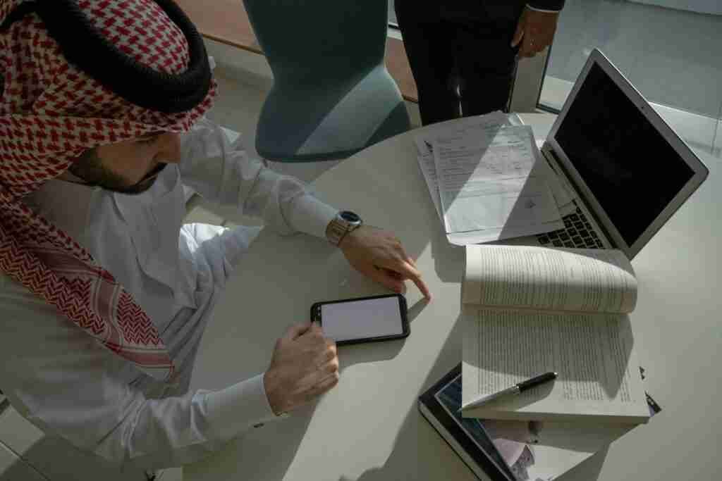 A man in traditional arab attire is focused on perusing phone contracts without credit checks on a tablet, with a laptop and an open book on the desk, suggesting a scene of study or business research.